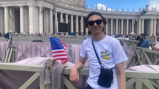 Brennan stands in front of the Vatican, a US stars and stripes flag is behind him