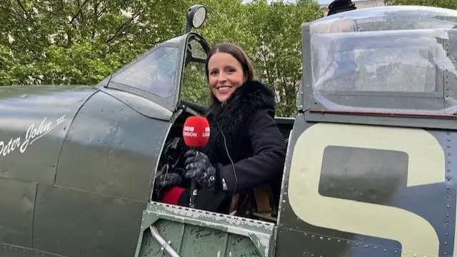 BBC reporter Gem O'Reilly (long brown hair, black coat with fur neck) holds a BBC-branded red mic while sitting in a Spitfire plane