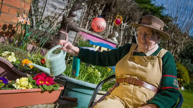 Annie outside, seated, watering some colourful flowers. She's wearing a brown hat, and khaki dungarees over a green jumper.