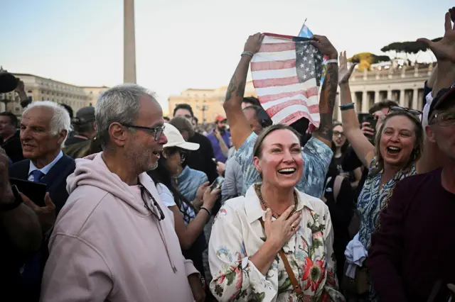 A woman holds her hand on her heart and smiles while another person holds up the US flag behind her in the crowd