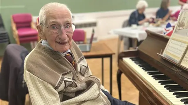 Stanley Carr smiles for the camera as he sits at a piano he wears glasses, a brown and white cardigan and a purple tie