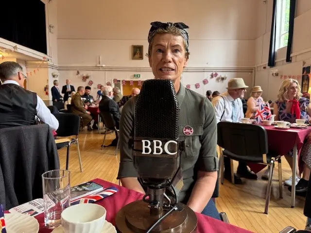 An old fashioned microphone on a stand with BBC in white letters stands on a table in front of Linda who is wearing a headscarf and an army green shirt