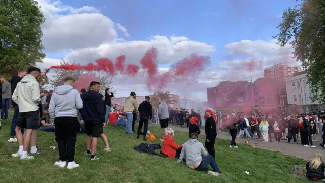 A large crowd of people gathering in the park. There are plumes of red smoke from flares drifting up into the air.