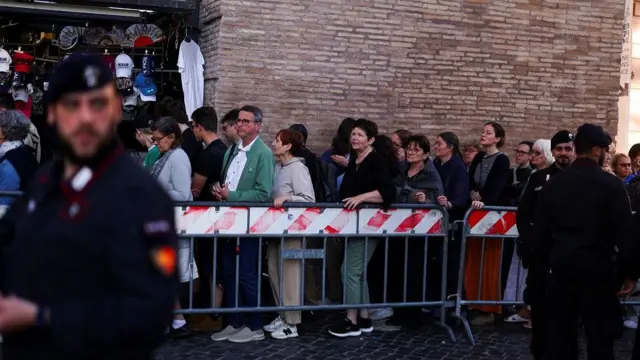A queue of people standing behind a metal barrier. Police officers stand in the foreground