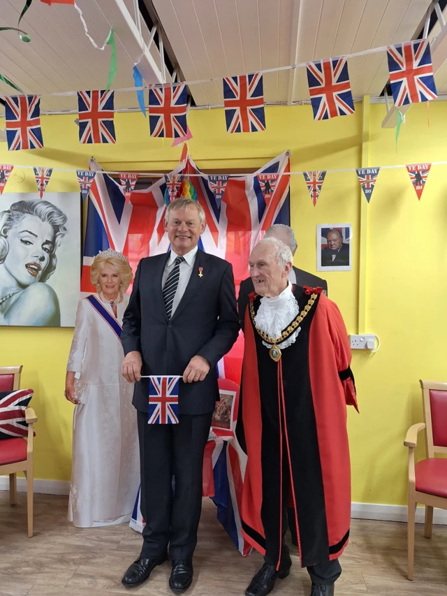 Martin Clunes standing up next to Terry Cordery, who is dressed in mayoral regalia, at the VE Day celebrations. They are stood in front of a cardboard cut-out of the Queen. On the wall there is a picture of Marilyn Monroe and Sir Winston Churchill. There is Union Flag bunting hung up around the room.