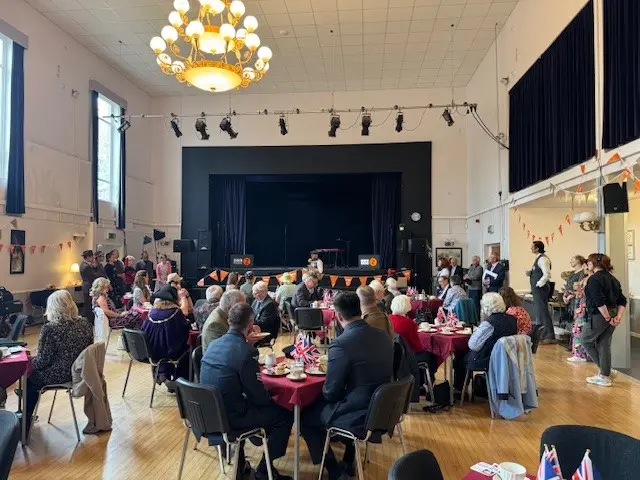 A crowd inside a large hall. A large stage is at the end of the room and features BBC Radio 2 branding. The audience are sitting at tables covered with burgundy table cloths and have a range of china tea cups, flag decorations and scones in front of them.