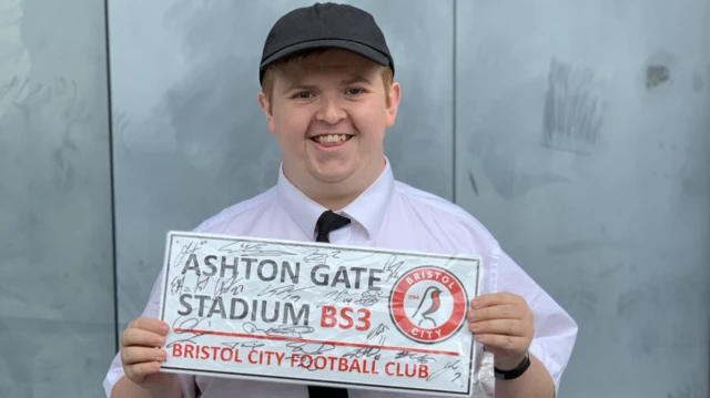Jack Beese wearing a white collared shirt with a black tie and a black cap.  He is holding up a sign which says Ashton Gate Stadium, Bristol City Football Club. It is covered in scrawled signatures.