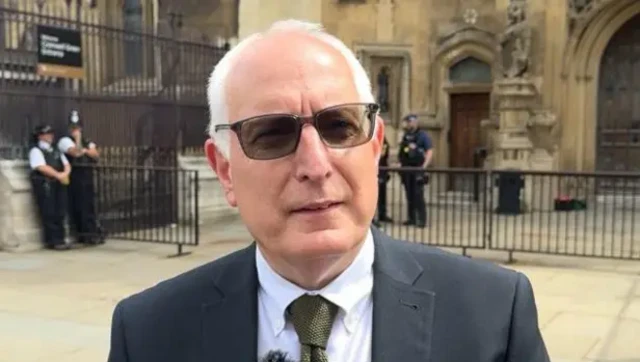 A picture of Neil Duncan-Jordan, who is wearing a grey jacket, green tie and white shirt. He has short white hair and is wearing sunglasses.