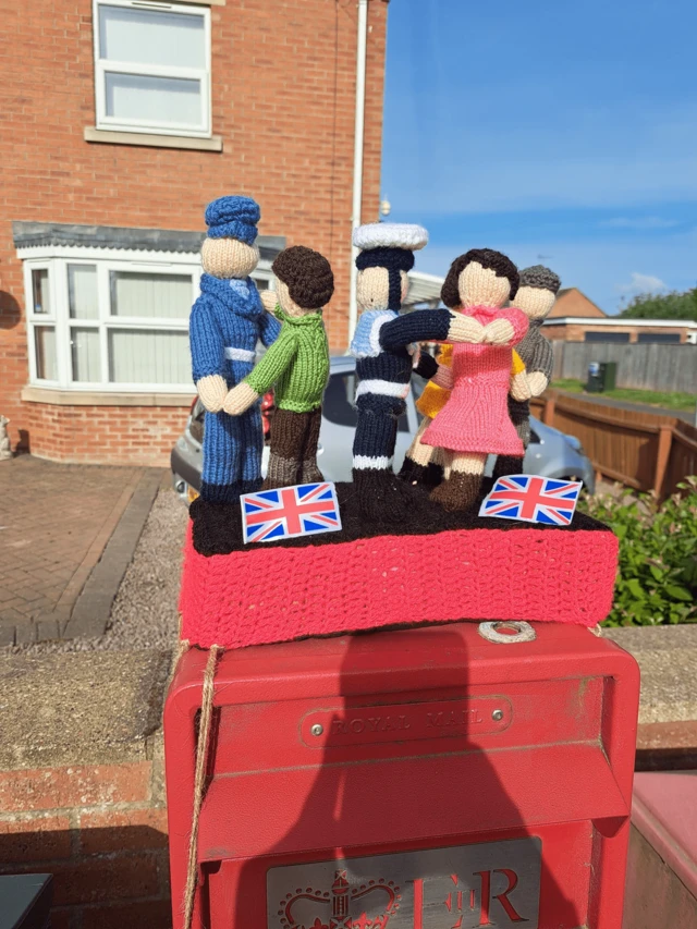 A square, red post box with a knitted scene on top. Six knitted figures are holding hands and look like they are dancing in scenes reminiscent of 1945.