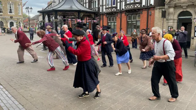 A group of swing dancers show off their moves in Brigg. Behind them is a Tudor-style wood and brick building and a gazebo decorated with Union flag bunting