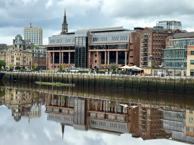 The imposing Newcastle Crown Court building made from red stone and large windows reflected in the River Tyne