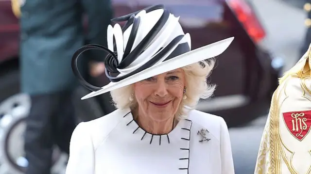 Queen Camilla in white morning coat dress with matching white and black hat. On her left lapel is a brooch of the 12th Lancers