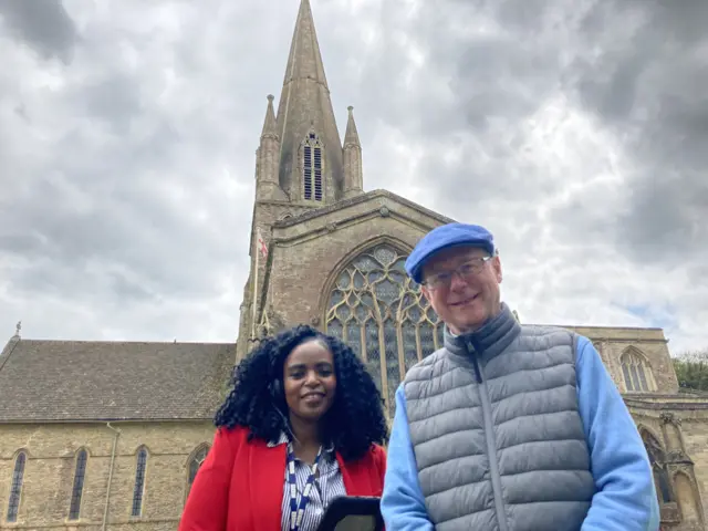 Bridgitte Tetteh, wearing a red jacket and blue and white shirt, standing next to Richard Young, who is wearing a blue jumper and flat cap. They are standing in front of St Mary's Church in Witney