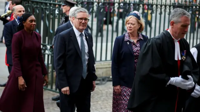 Keir Starmer in a balck suit arrives at Westminster Abbey, Conservative leader Kemi Badenoch in a purple morning coat walking behind her