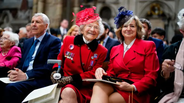 Two women wearing red jackets, one with veterans badges, and both wearing fancy hats, smile at the camera while sitting inside the abbey