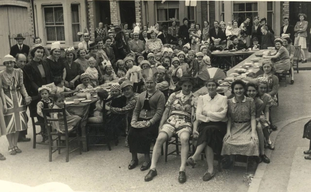 A black and white picture of people sat around tables celebrating with end of World War Two, with some in fancy dress.