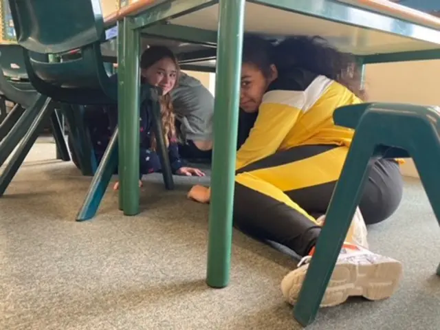 Children taking part in a mock air raid at a school in Hull. They are under a desk and looking at the camera.