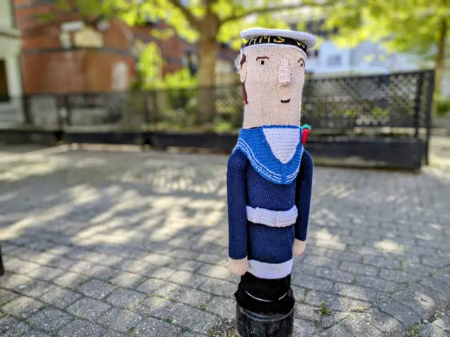 A woollen sailor placed over a bollard. He is wearing a blue sailor outfit and a poppy.