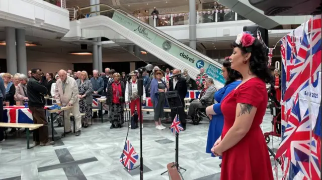 Woman in red dress stands in front of microphone on bottom floor of shopping centre, silent, whilst crowd stands, also observing silence.