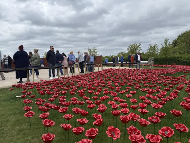 Visitors to the International Bomber Command Centre stand on a gravel path behind a wooden fence and look out at a grass field covered in red ceramic poppies. The field is surrounded by green bushes and trees. The sky is grey and cloudy.