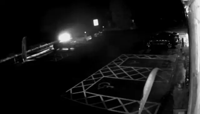 Still from black and white CCTV showing a dark car park and a pair of headlights passing on a road nearby