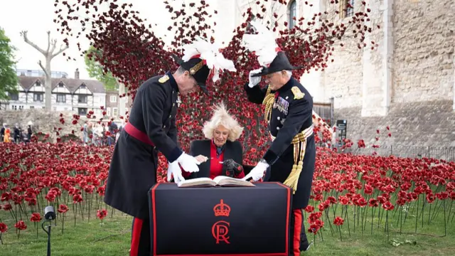 Queen Camilla signing a visitors book in front of an explosion of ceramic poppies.