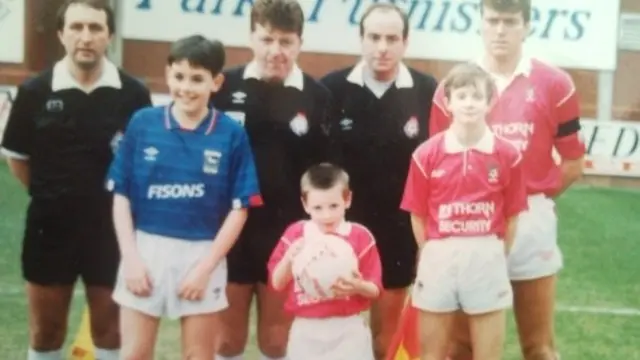 An old group picture showing Dave Tovey as a young boy. He is wearing a red football shirt and white shorts, holding a football and kneeling down on the pitch bedside other players.
