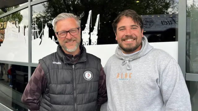 Barry wearing a black Bristol City gilet and black glasses. He is standing beside Nathan, who is wearing a grey hoodie. They are both smiling at the camera.