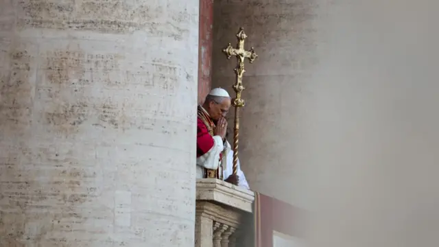 The pope bows his head on the balcony and holds his hands together in prayer, with a large cross held up behind him