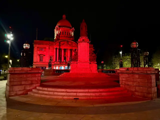 Red light illuminates a statue of Queen Victoria which stands on top of a plinth in the city centre. The Hull City Hall, which is also lit up with red light, is visible in the background.