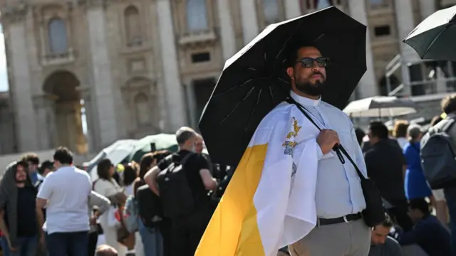 A priest holding a black umbrella and wearing a Vatican City flag draped over his right shoulder