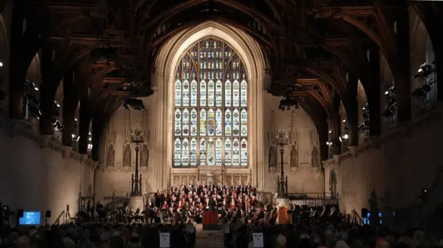 view from the back of the hall, of a choir performing on the stage of a grand gothic hall, with audience watching.