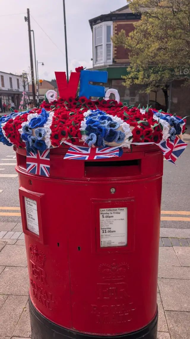 A large red, white and blue knitted post box topper recreating a union flag design in small poppies. It also features the word VE 80 in the centre.