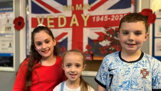 Three smiling primary-aged children stand in front of a Union Flag that reads "8th may, VE DAY"