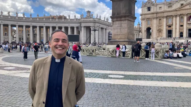 Pedro Manuel wearing a tan jacket and trousers, as well as the black shirt and dog collar. He is staning in St Peter's Square and smiling broadly.