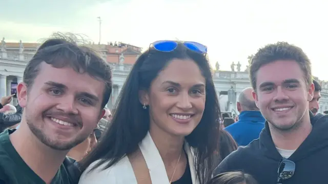 Lissette smiles at the camera while stood in St Peter's Square with two men who are also smiling at the camera. She has brown hair and eyes and sunglass resting on top of her head.