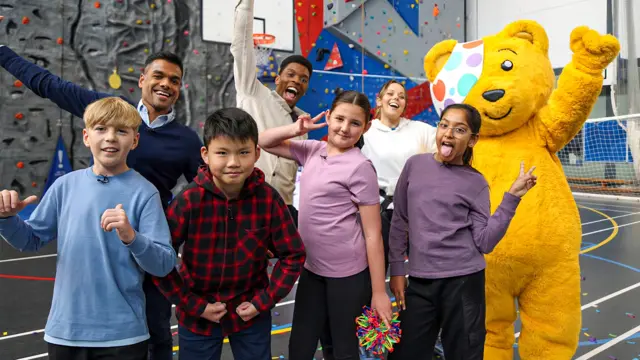 Presenters, Pudsey Bear and children posing in front of a climbing wall.