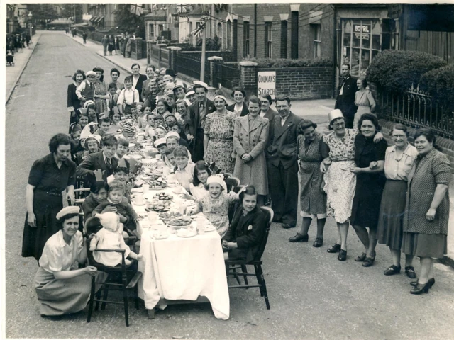 A black and white picture showing residents in Reading standing for a picture. Children are sat at a long table which is decorated with a white cloth, with adults stood up around it and smiling for the camera.