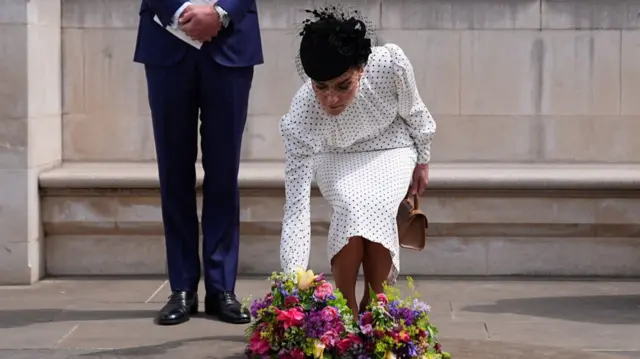 The Princess of Wales lays flowers at the Innocent Victims' Memorial in a white polka dot dress with black hat with veil. to her right is Prince William, visible from the mid-rift down