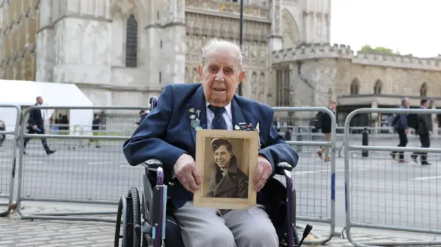 An elderly man on a wheelchair poses in front of Westminster Abbey holding a picture of himself when he was younger