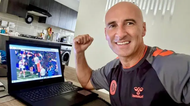 Dean Allen wearing a football shirt and sitting at a dining table in front of his laptop, which is playing a football match. He is bald and is smiling at the camera with a fist in the air.