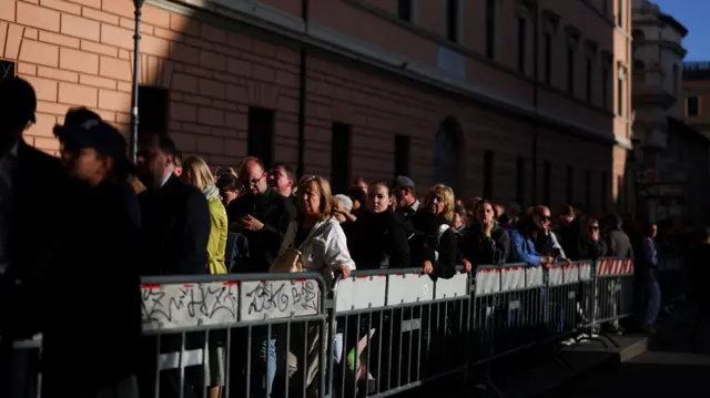 A line of people queue up behind a metal barrier