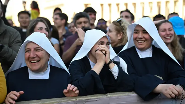 Three nuns wearing white head dresses smile and cheer in Vatican Square