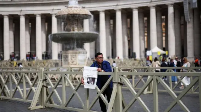 A man leaning over a fence takes a photo of himself holding Vatican newspaper L'Osservatore Romano