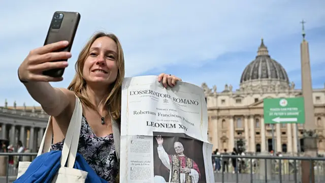 A woman poses, uses her smartphone and holds a newspaper with the picture of newly elected pope Leo XIV, with St Peter's Basilica in the background in The Vatican