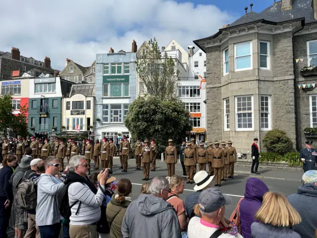A crowd of people line a road, which has people in military uniforms standing to attention. Behind there are tall granite buildings.