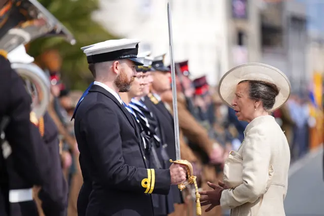A grey haired woman in a cream dress and hat speaks to a man in a blue naval jacket, with a white cap who is holding a sword.