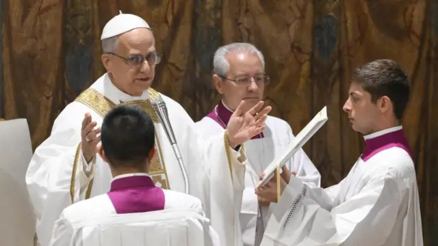 Pope Leo speaking into a microphone, two alter boys are in front of him, one holding a document for him to read from,and a priest behind him