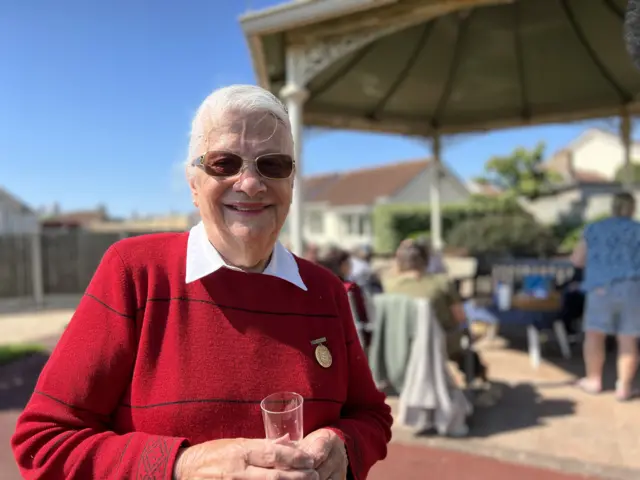A lady is pictured wearing a red shirt and holding a glass. She has grey hair and is wearing black glasses, while smiling at the camera