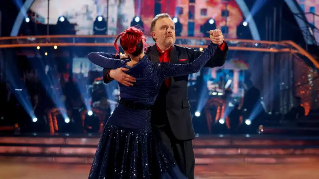 Diane Buswell and Chris McCausland dancing - he is wearing a red shirt and dark suit, she is wearing a navy sequinned dress.